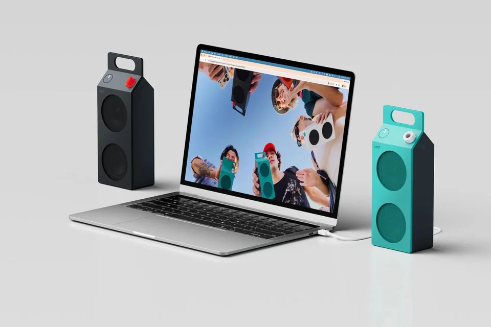Zwei Lautsprecher in Dunkelgrau und Türkis neben einem Laptop, einer per USB-C-Kabel verbunden