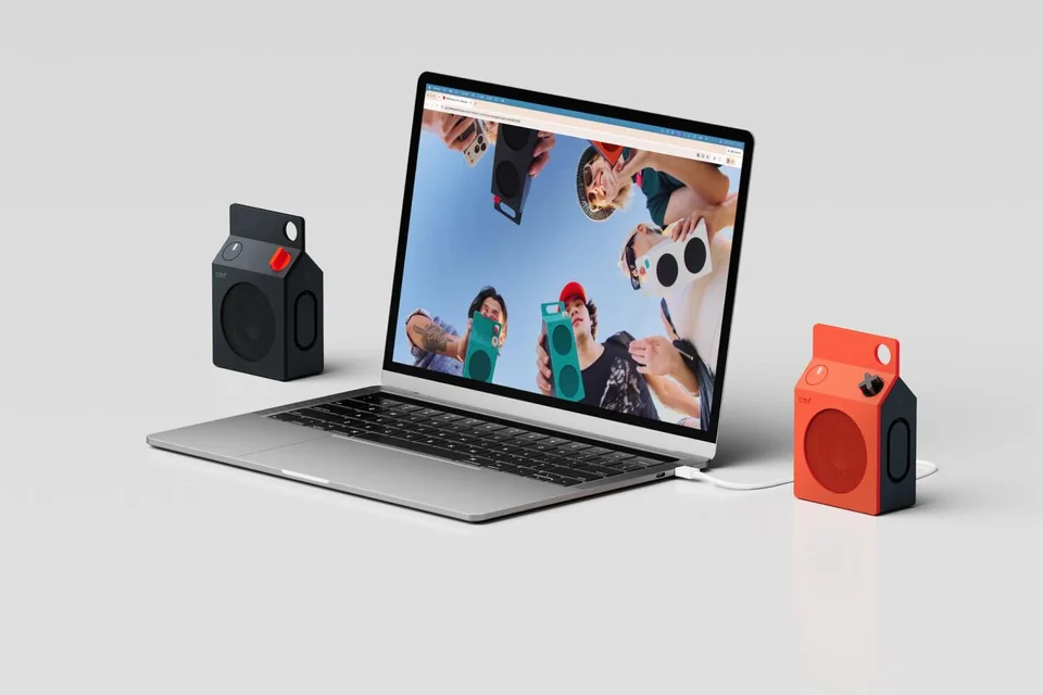 Zwei kompakte Lautsprecher in Dunkelgrau und Orange neben einem Laptop, einer per USB-C-Kabel angeschlossen