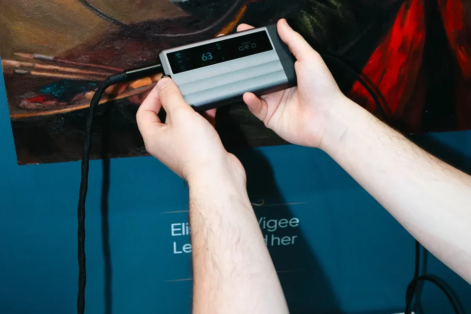 Zwei Hände halten einen mobilen DAC-Kopfhörerverstärker mit angeschlossenem Kabel vor einem Gemälde