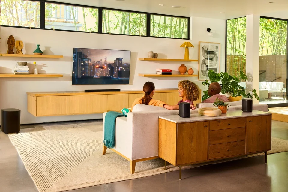 Wohnzimmer mit Soundbar unter dem Fernseher, Subwoofer und zwei schwarzen Lautsprechern hinter dem Sofa