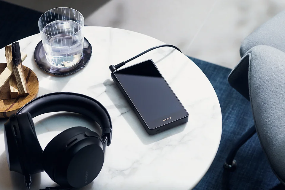 Schwarzer Walkman mit angeschlossenem Kabel auf einem weißen Marmortisch neben Over-Ear-Kopfhörer und Wasserglas